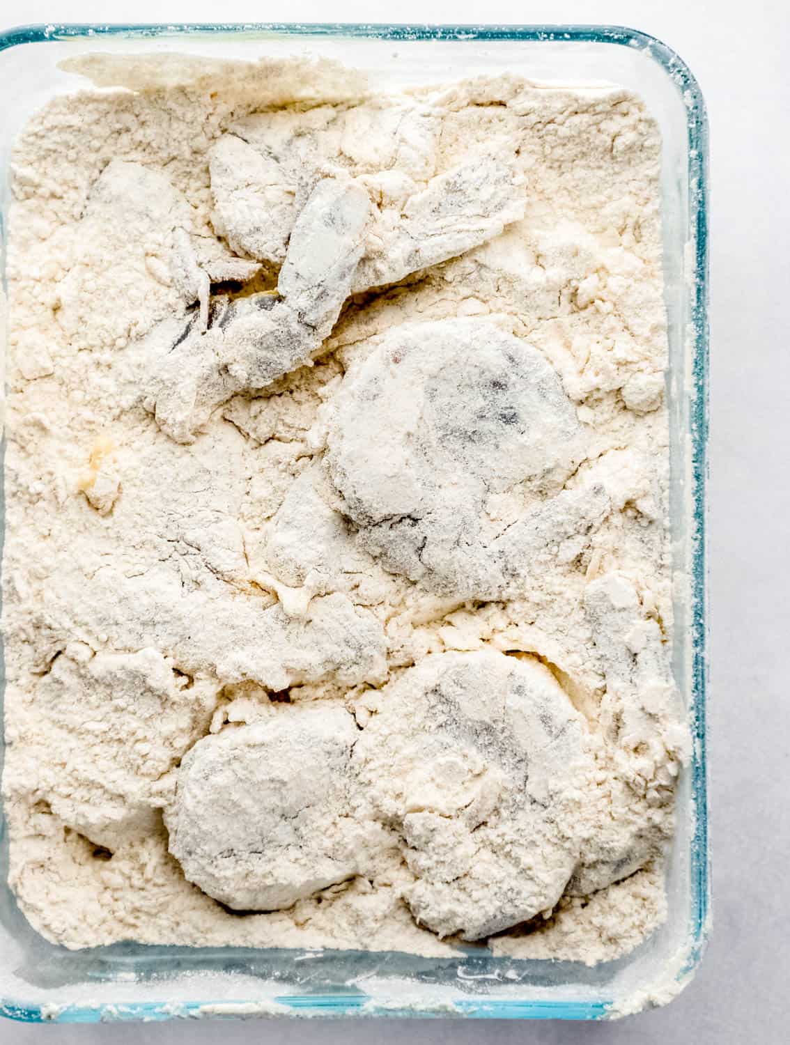 Overhead view of shrimp in flour mixture inside a rectangle glass container. 