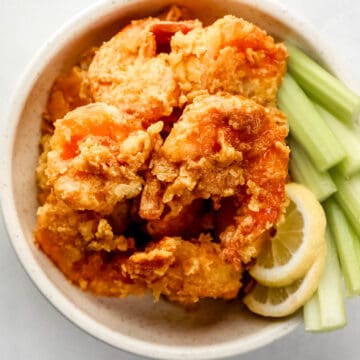 Overhead view of buffalo shrimp in white bowl with celery sticks and lemon slices.