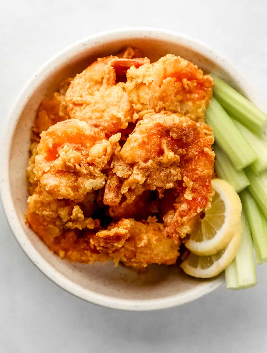 Overhead view of buffalo shrimp in white bowl with celery stick and lemon slices. 