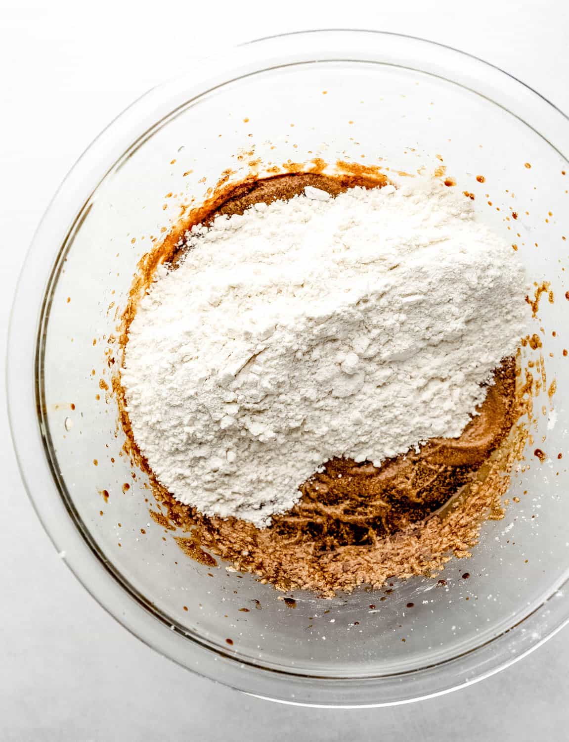 Overhead view of flour mixture added to wet cookie ingredients in large glass mixing bowl. 