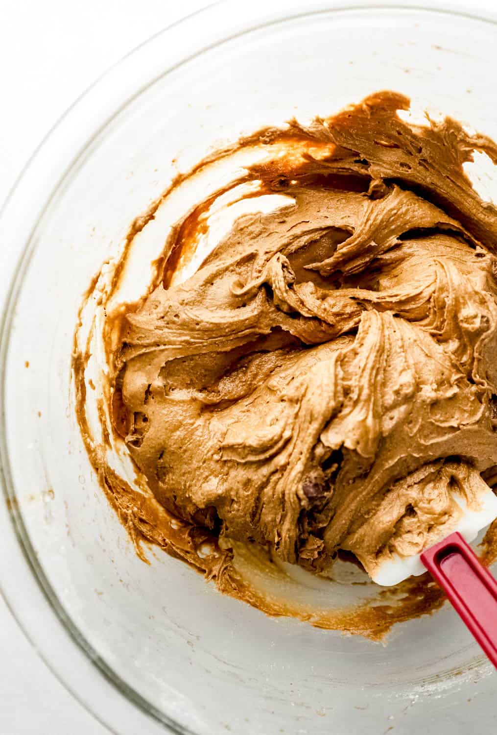 Chocolate chunks being folded into cookie dough in large glass mixing bowl with spatula. 