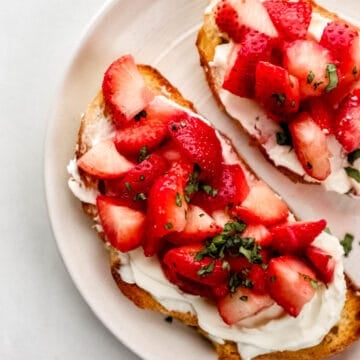 Overhead view of two slices of cream cheese toast topped with strawberries and basil on white plate.