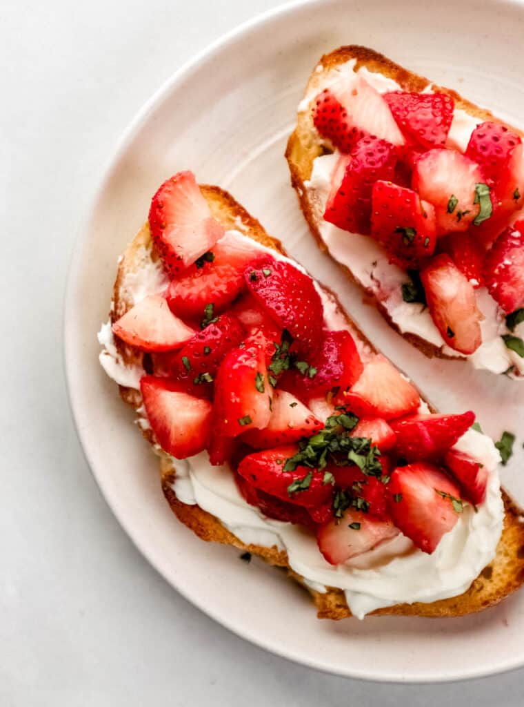 Overhead view of two slices of cream cheese toast topped with strawberries and basil on white plate.