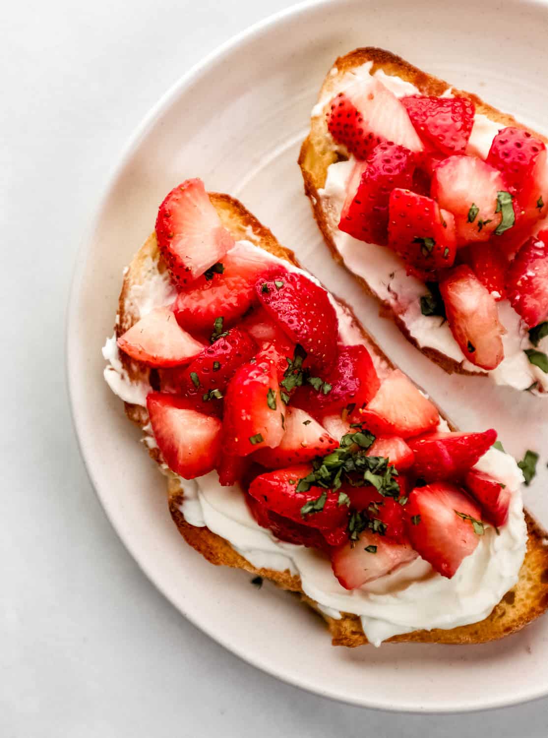 Overhead view of cream cheese toast topped with strawberries and basil on white plate 