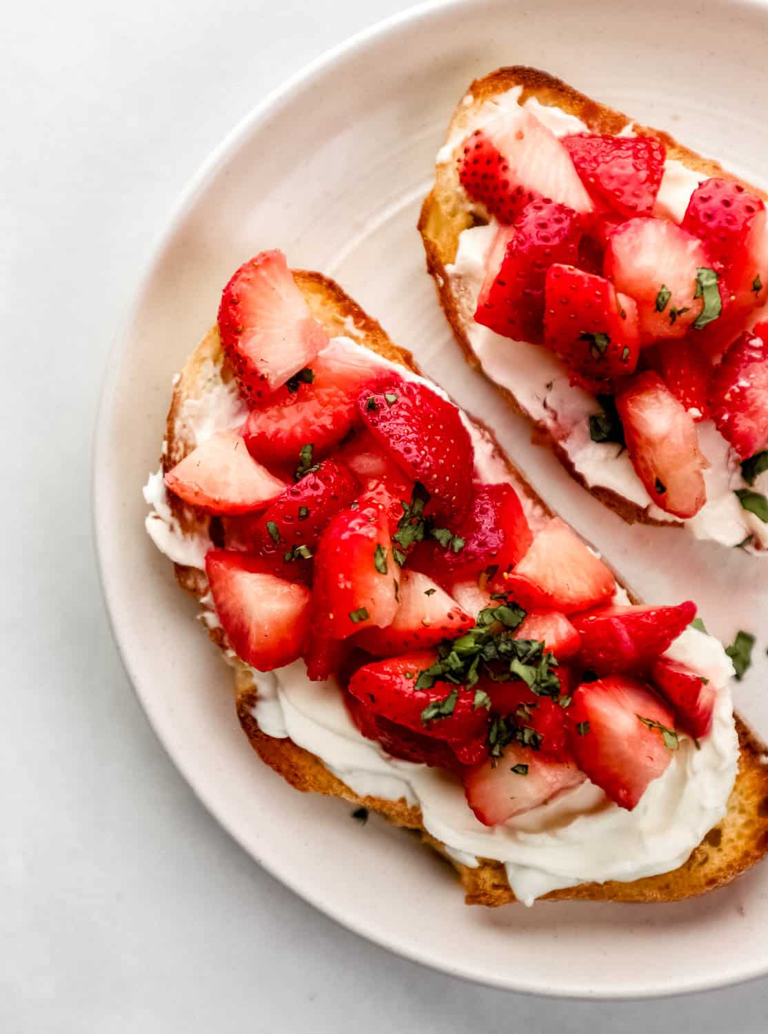 Overhead view of cream cheese toast topped with strawberries and basil on white plate