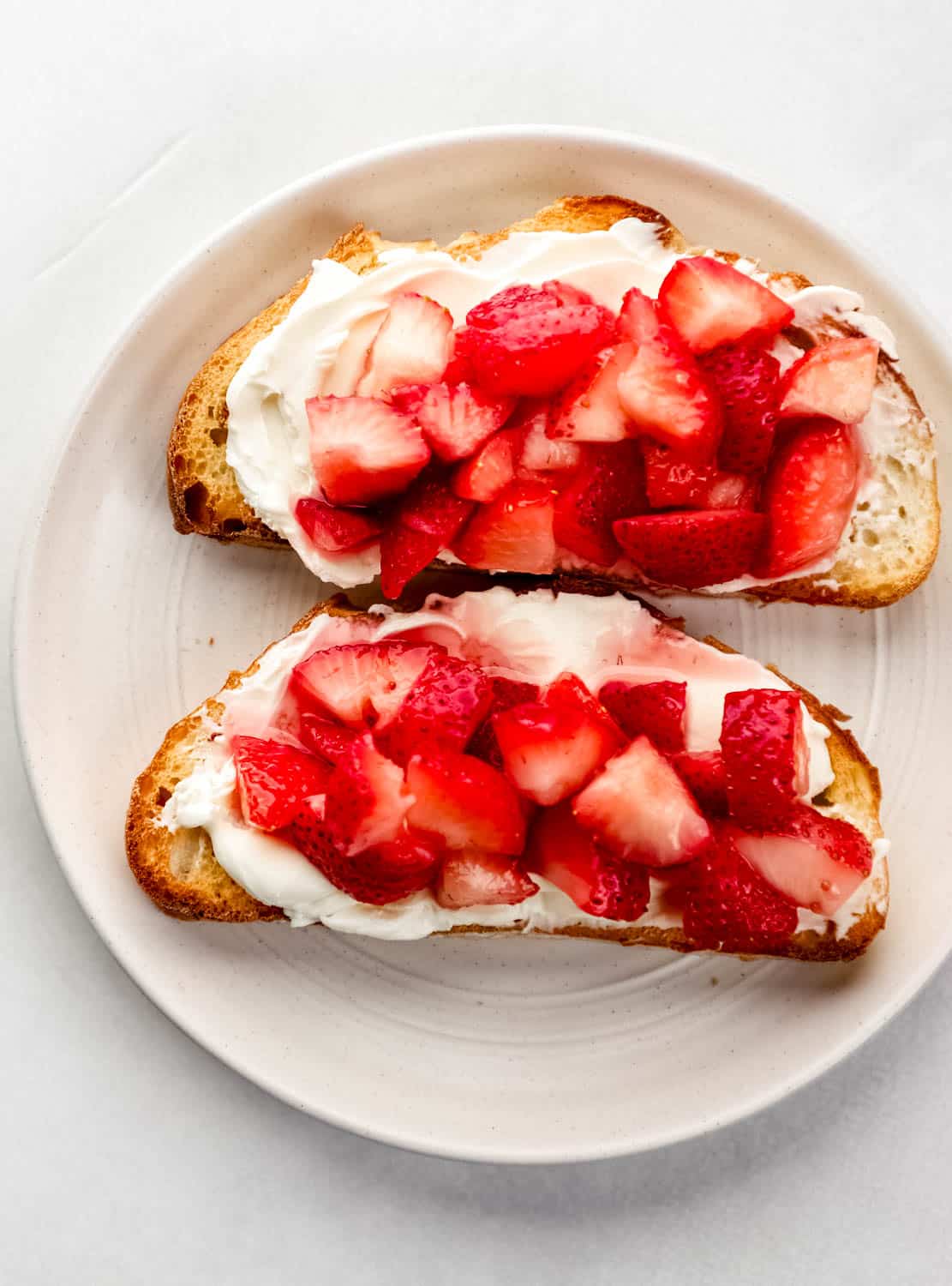 Two slices of cream cheese toast on white plate topped with strawberries. 