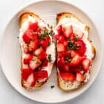 Overhead view of two slices of cream cheese toast topped with strawberries and basil on white plate.