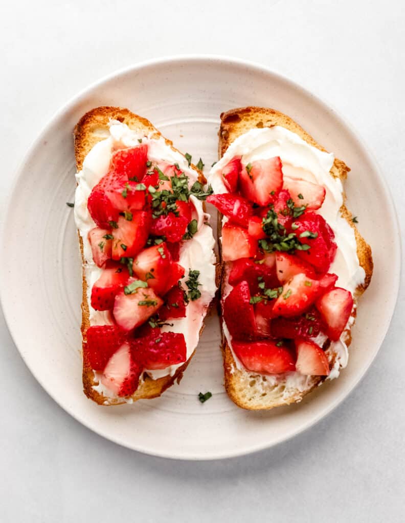 Overhead view of two slices of cream cheese toast topped with strawberries and basil on white plate.