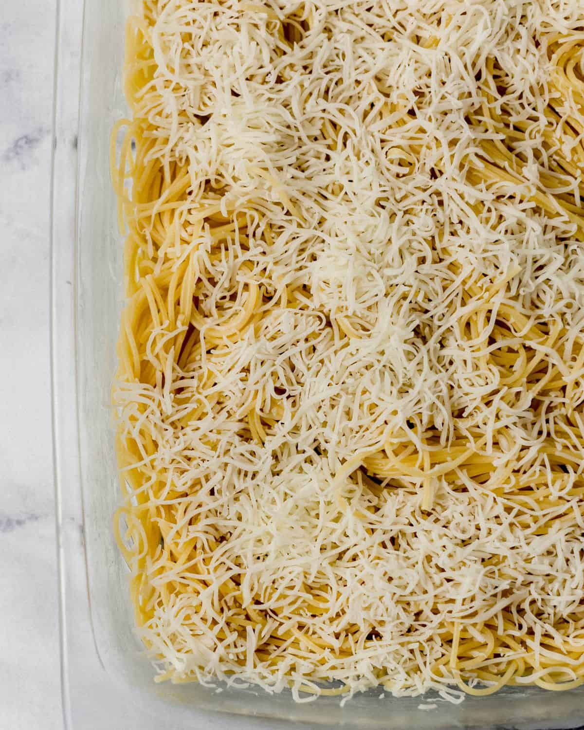 Overhead view of glass rectangle baking dish with cooked spaghetti and shredded cheese in it. 