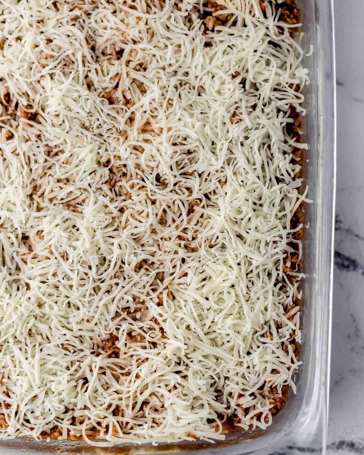 Meat sauce topped with shredded cheese in glass baking dish on marble surface. 