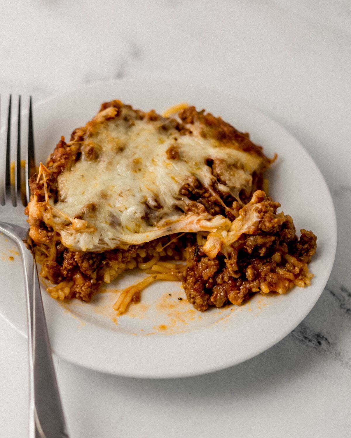 Baked spaghetti on white plate with fork in it on marble surface. 