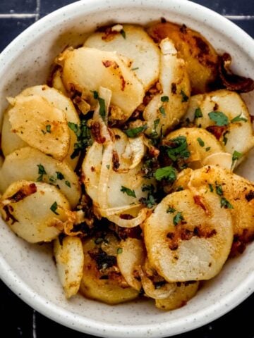 Overhead view of fried potatoes topped with fresh parsley in white bowl on black tile surface by a fork.