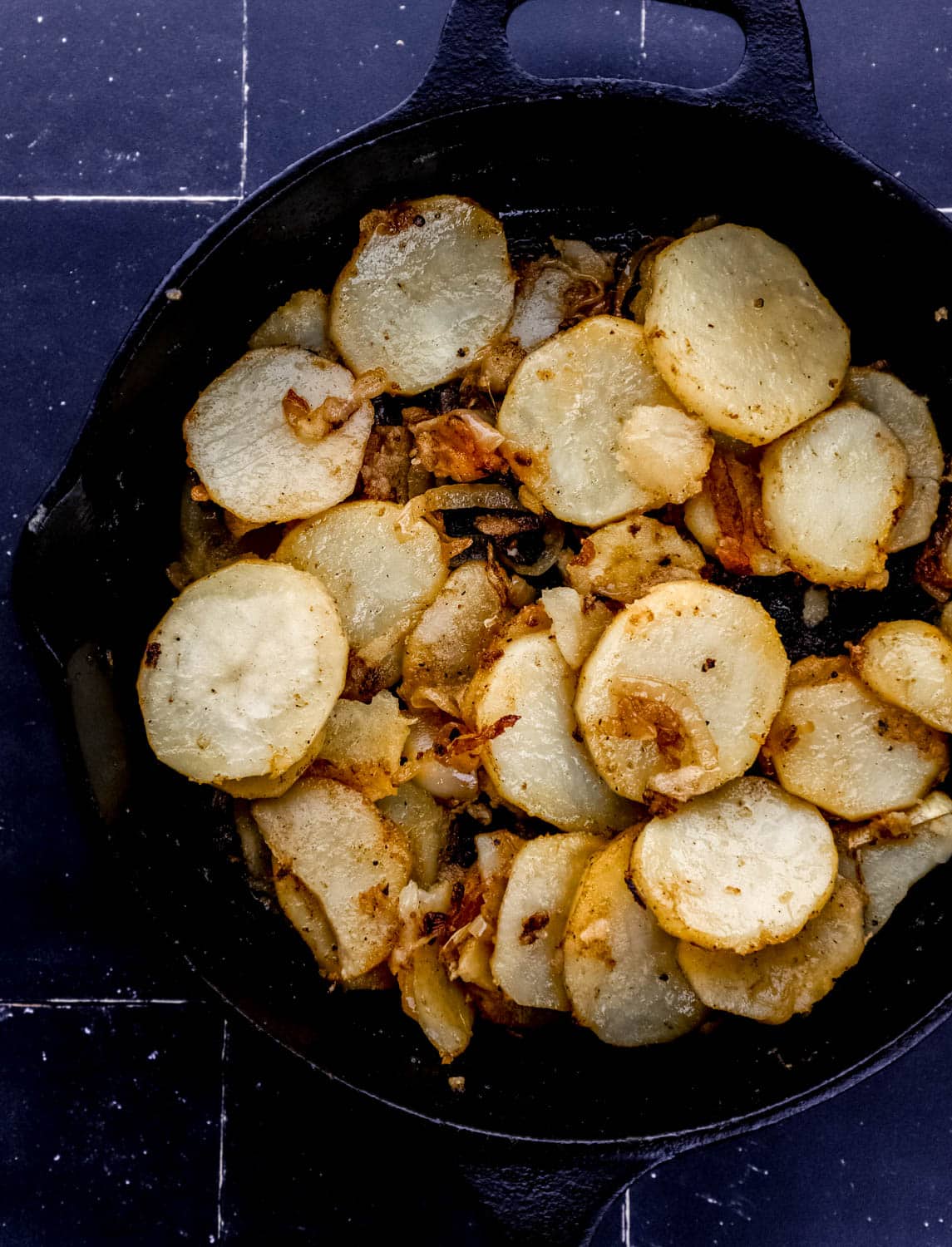 Overhead view of cooked potatoes in cast iron skillet on black tile surface. 