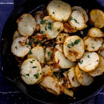 Overhead view of fried potatoes in cast iron skillet on black tile surface.