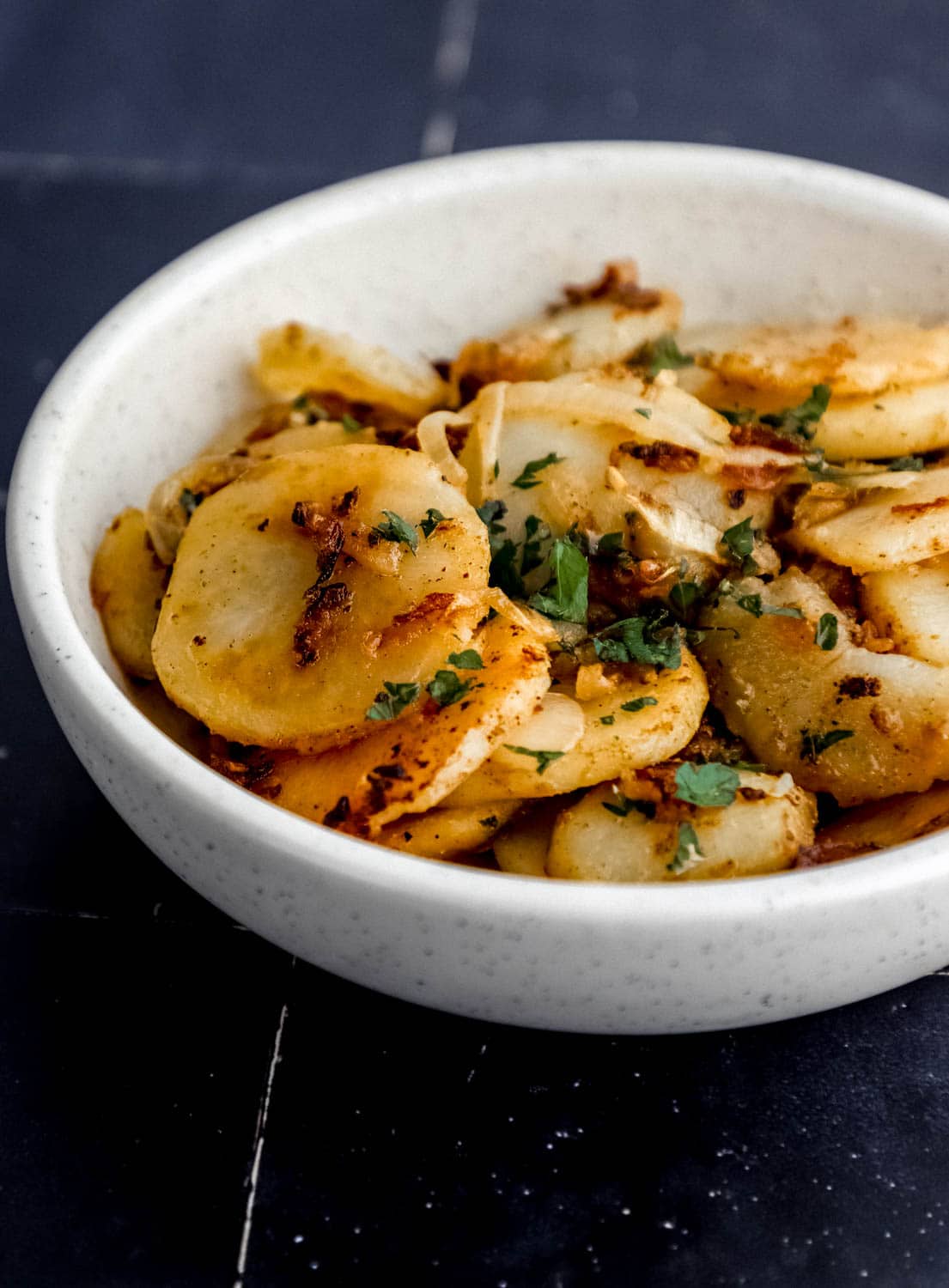 Close up side view of finished potatoes in a white bowl on black tile surface. 