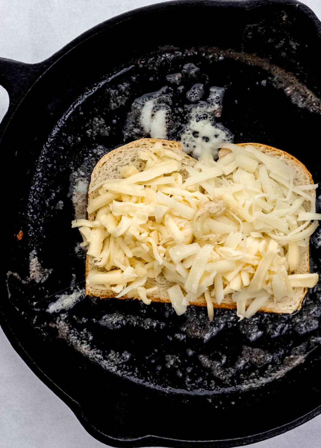Overhead view of slice of bread topped with shredded cheese in buttery cast iron skillet. 
