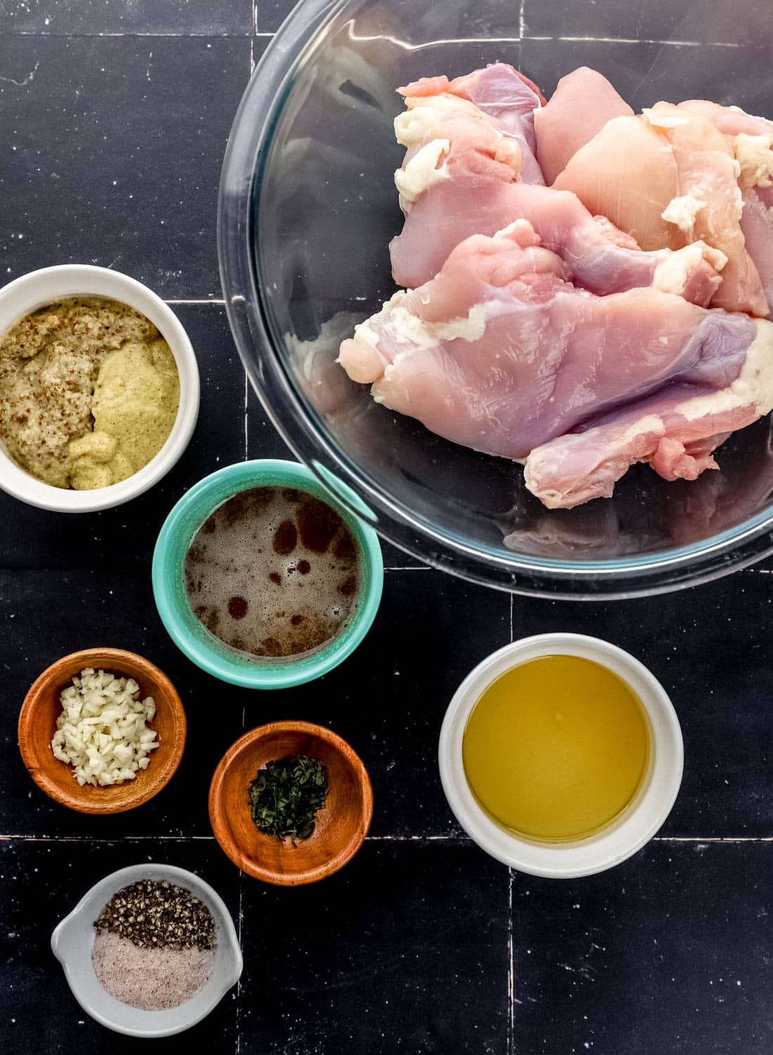 Overhead view of ingredients needed to make chicken in separate bowls on black tile surface. 