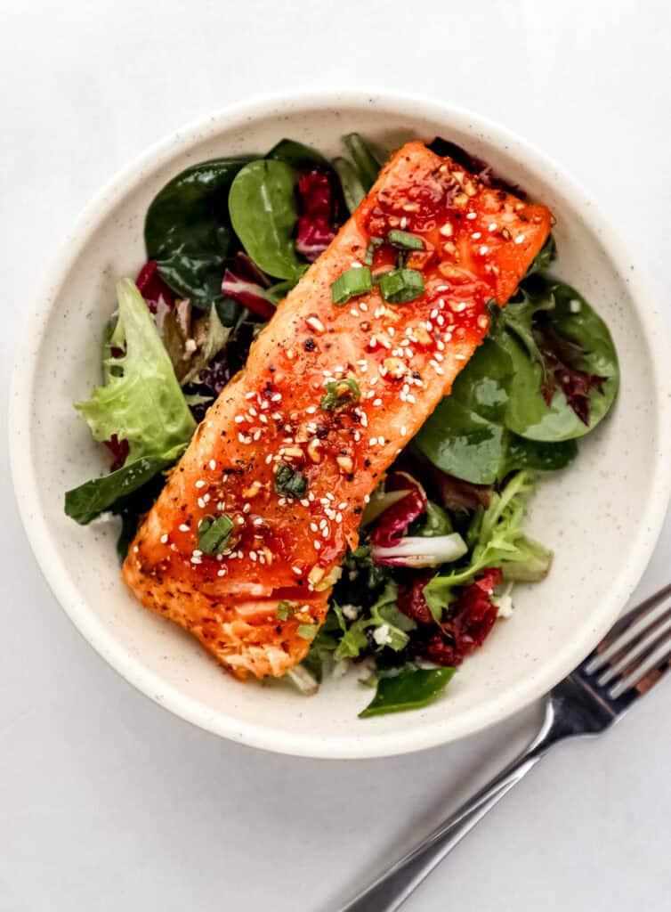 Overhead view of sriracha salmon on top of salad greens in a white bowl beside a fork.