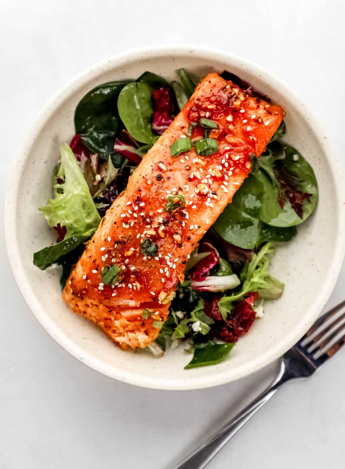 Overhead view of white bowl with finished salmon on top of salad beside a fork on white surface. 