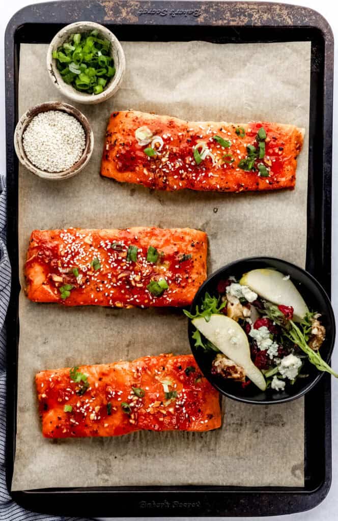Overhead view of baked salmon on parchment lined baking sheet with three bowls on it with sesame seeds, green onion, and salad in it.