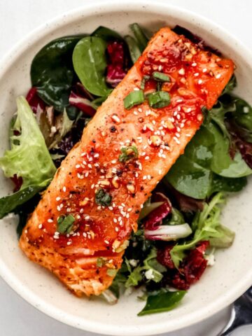 Overhead view of white bowl with salmon on top of salad beside a fork on white surface.