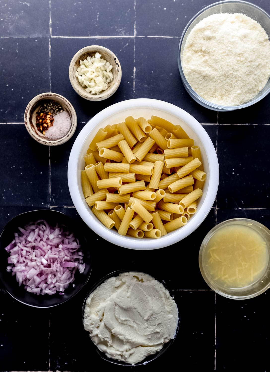 Overhead view of ingredients needed to make ricotta pasta in separate bowls on black tile surface. 
