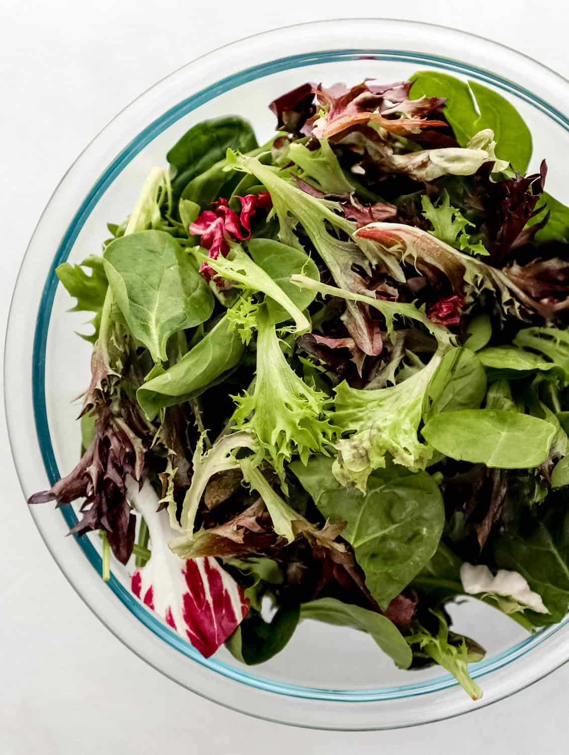 Mixed salad greens added to large glass mixing bowl. 