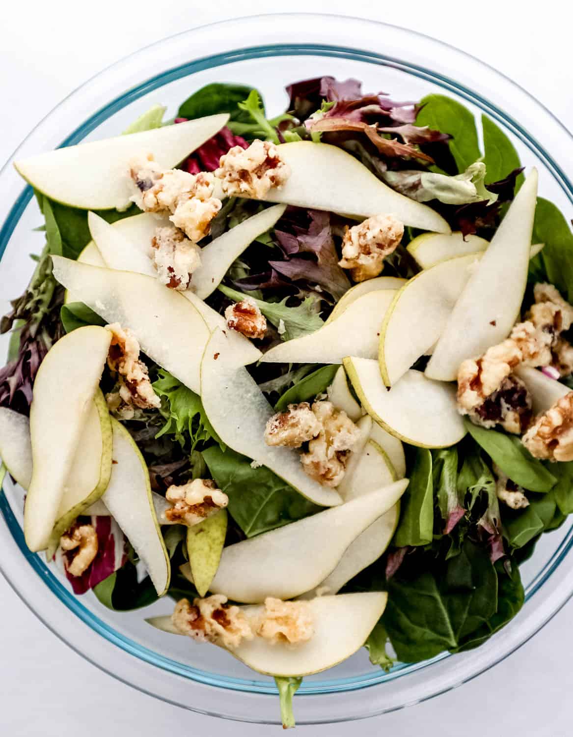 Large glass mixing bowl with salad greens, pear slices, and candied walnuts in it. 