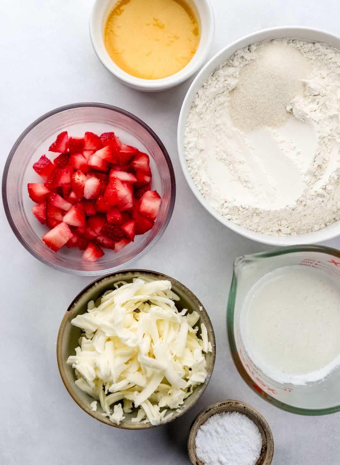 Overhead view of ingredients needed to make biscuits in separate bowls on white surface. 