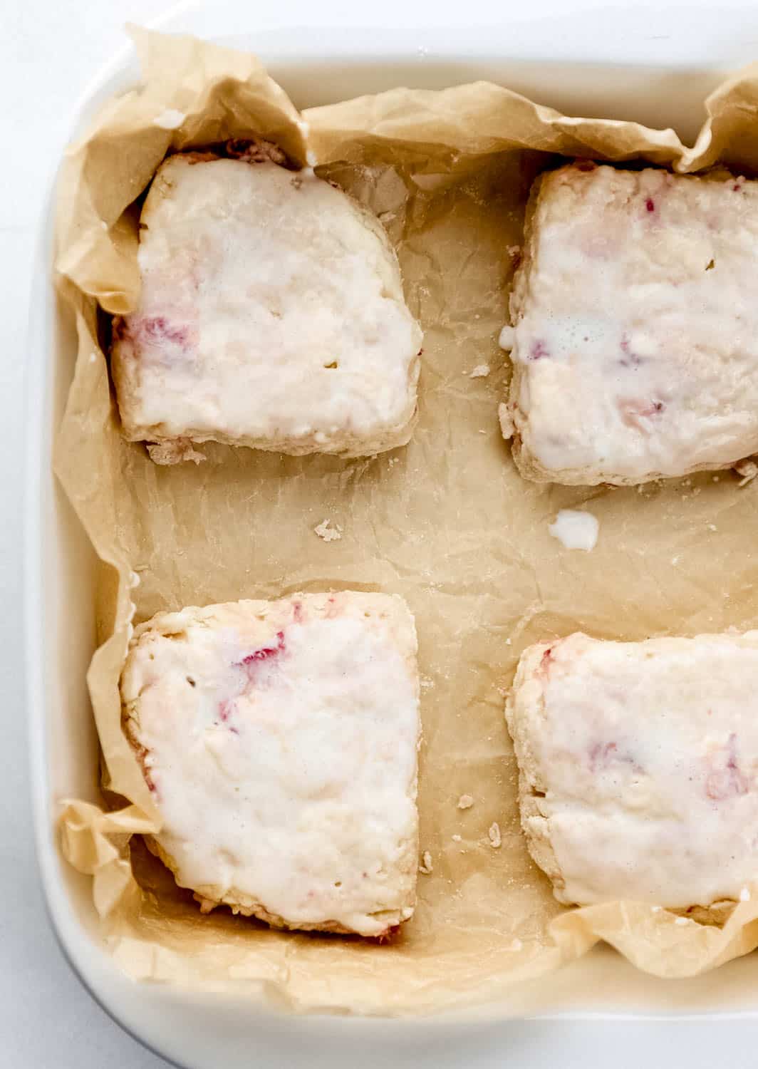 Overhead view of biscuits brushed with buttermilk in parchment lined baking dish. 