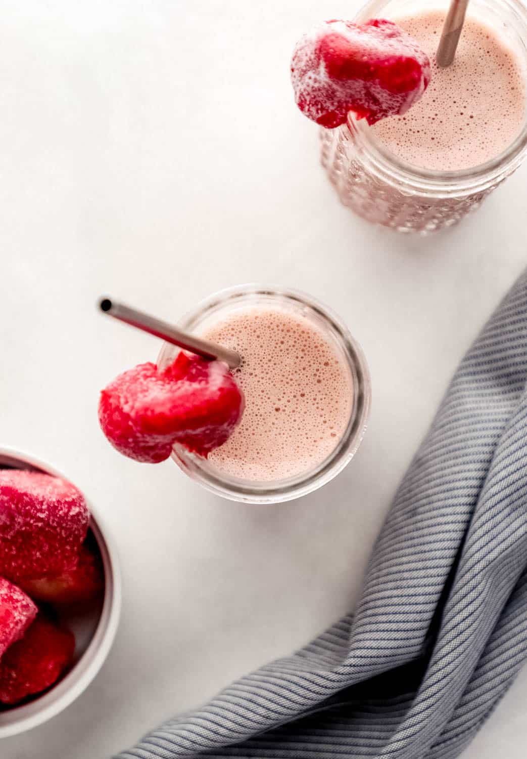 Overhead view of two smoothies in glasses with straws by a cloth napkin and bowl of frozen strawberries. 