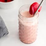 Close up side view of strawberry smoothie in a glass with straw in it and strawberry on top of it.