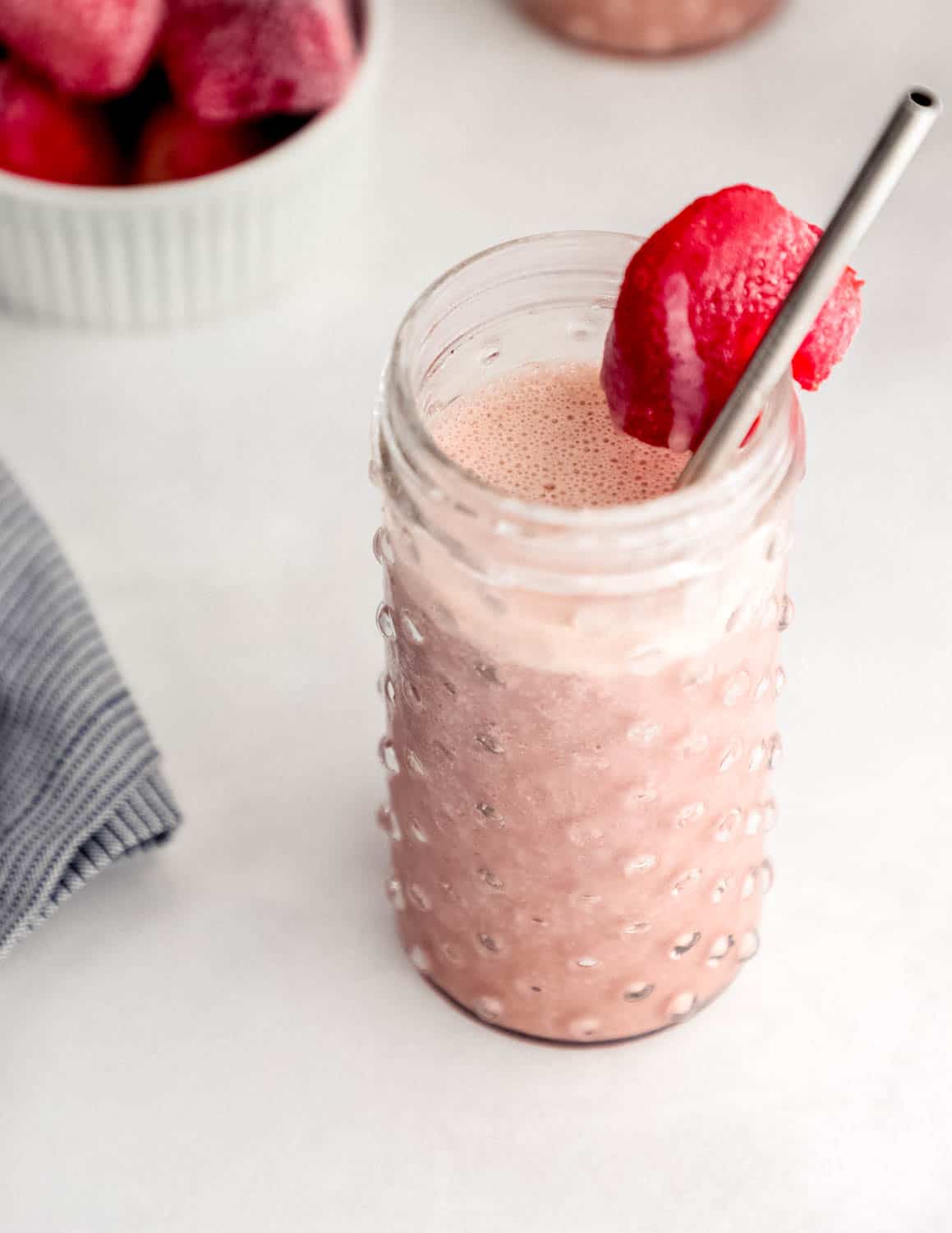 Close up side view of smoothie in glass with white bowl of frozen strawberries in the background. 