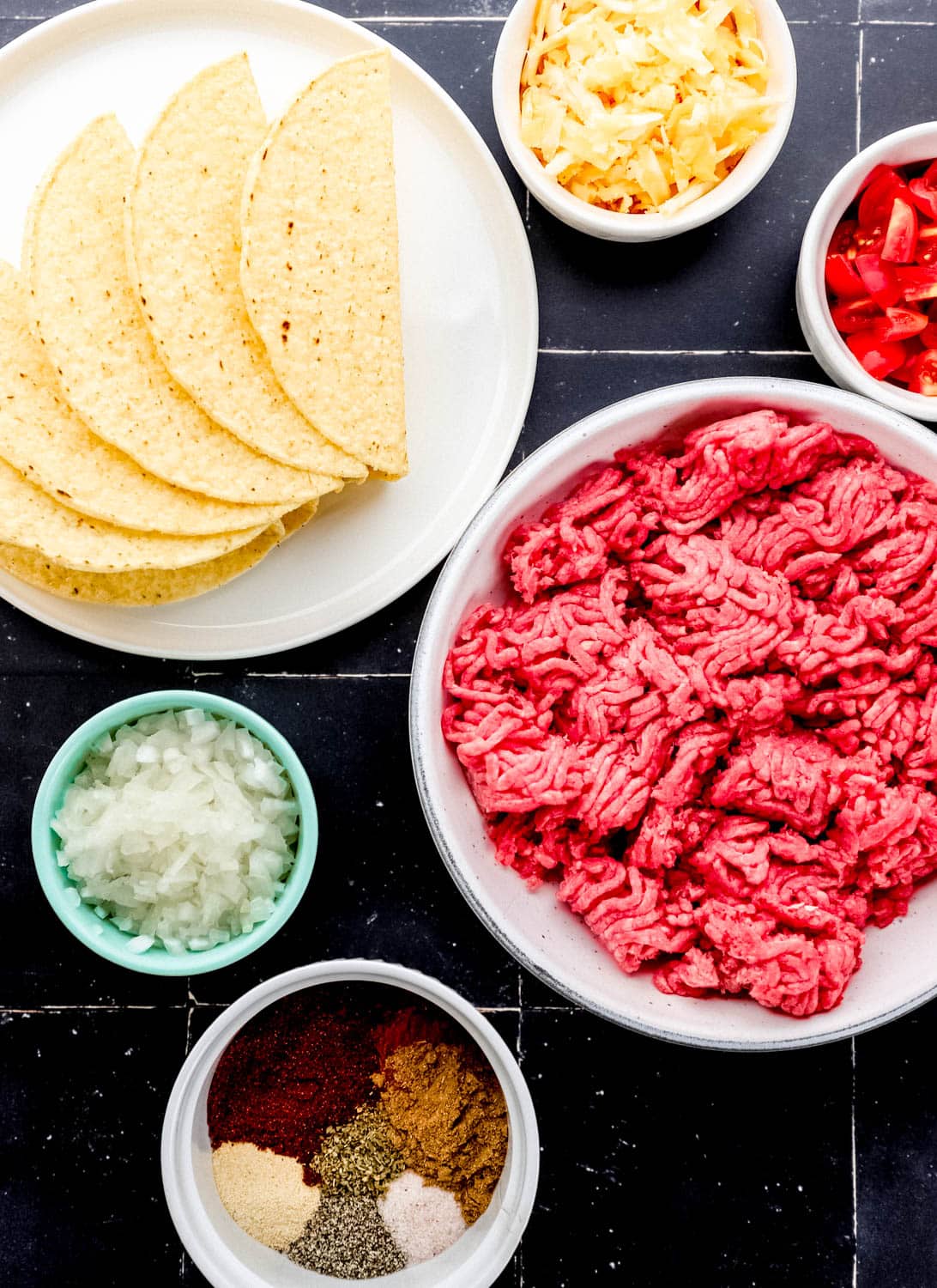 Overhead view of ingredients needed to make tacos in separate bowls on black tile surface. 