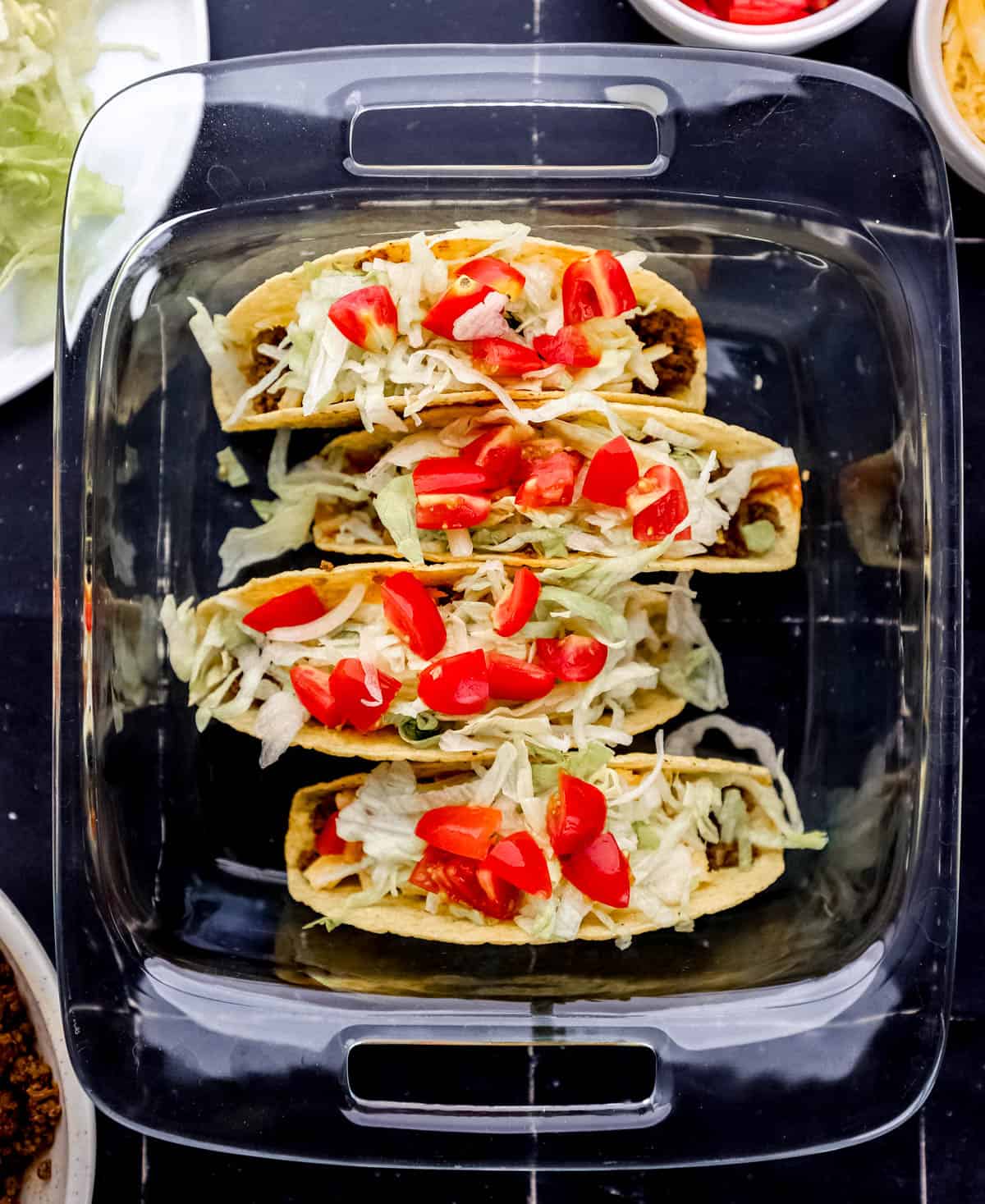 Tacos topped with lettuce and tomato in glass baking dish. 