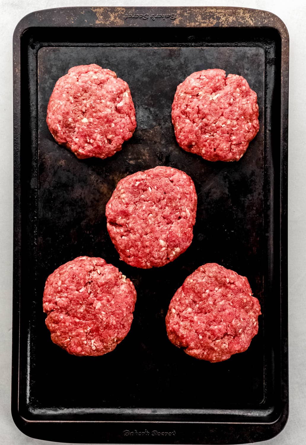 Overhead view of raw hamburger patties on sheet pan. 