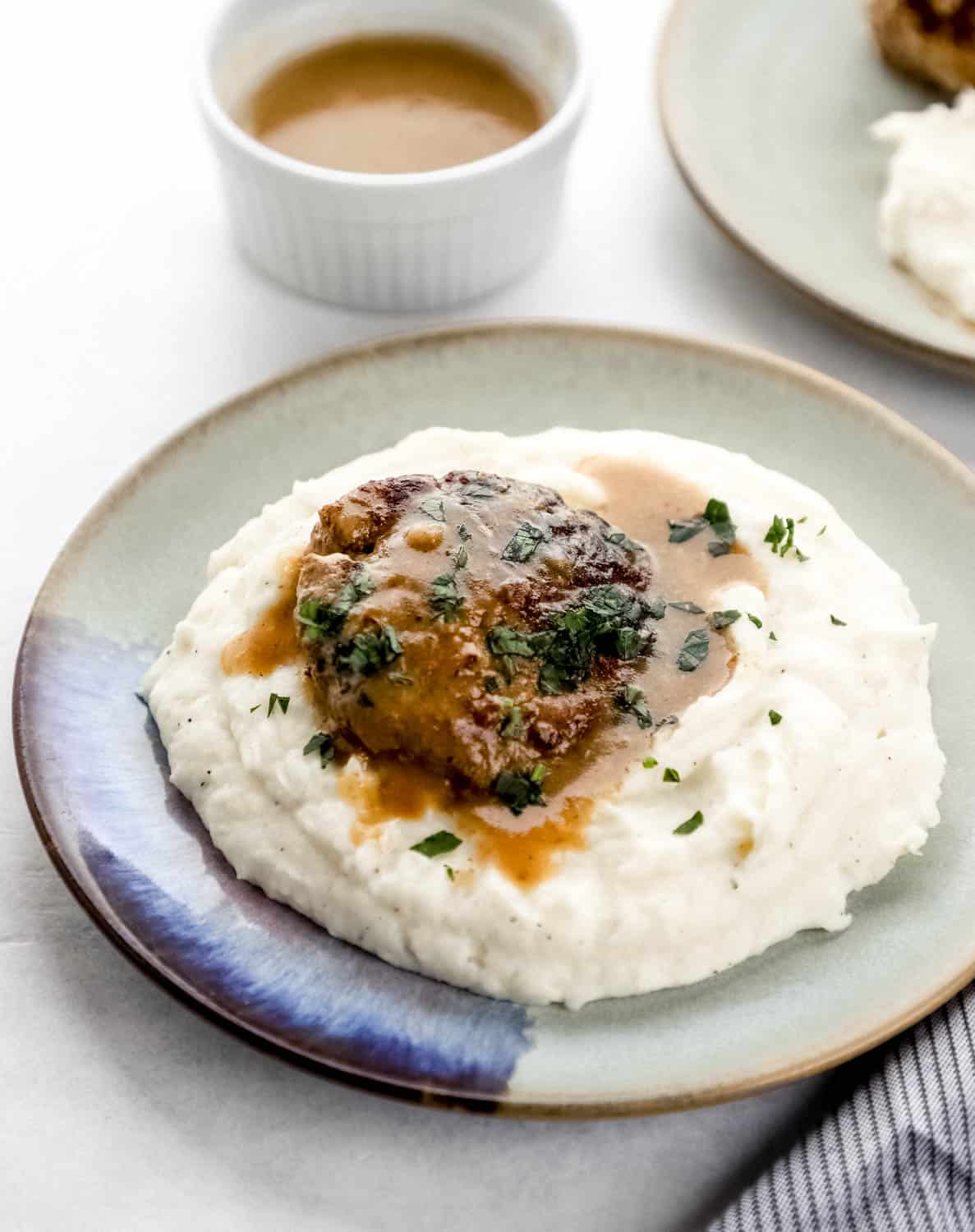 Close up side view of steak, gravy, and mashed potatoes on a plate with small white bowl of gravy in the background. 