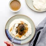 Overhead view of plates with mashed potatoes, burger and gravy on it by cloth napkin and small white bowl of gravy.