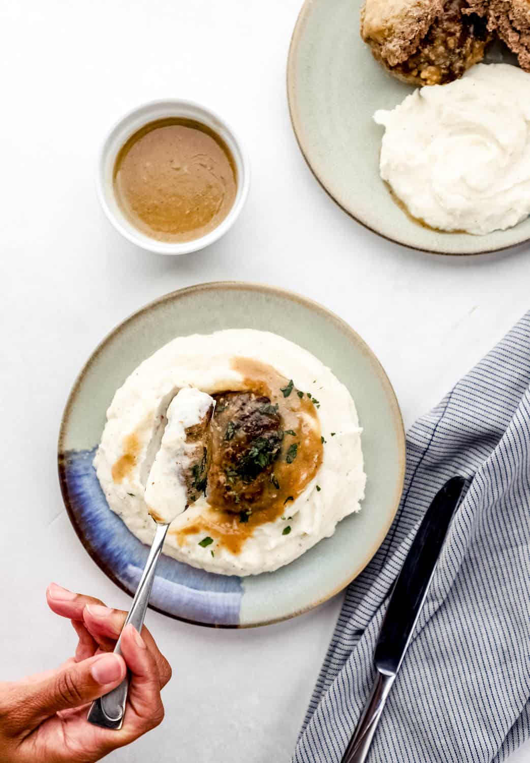 Overhead view of burgers, gravy, and mashed potatoes on plates by a cloth napkin and small bowl of gravy. 