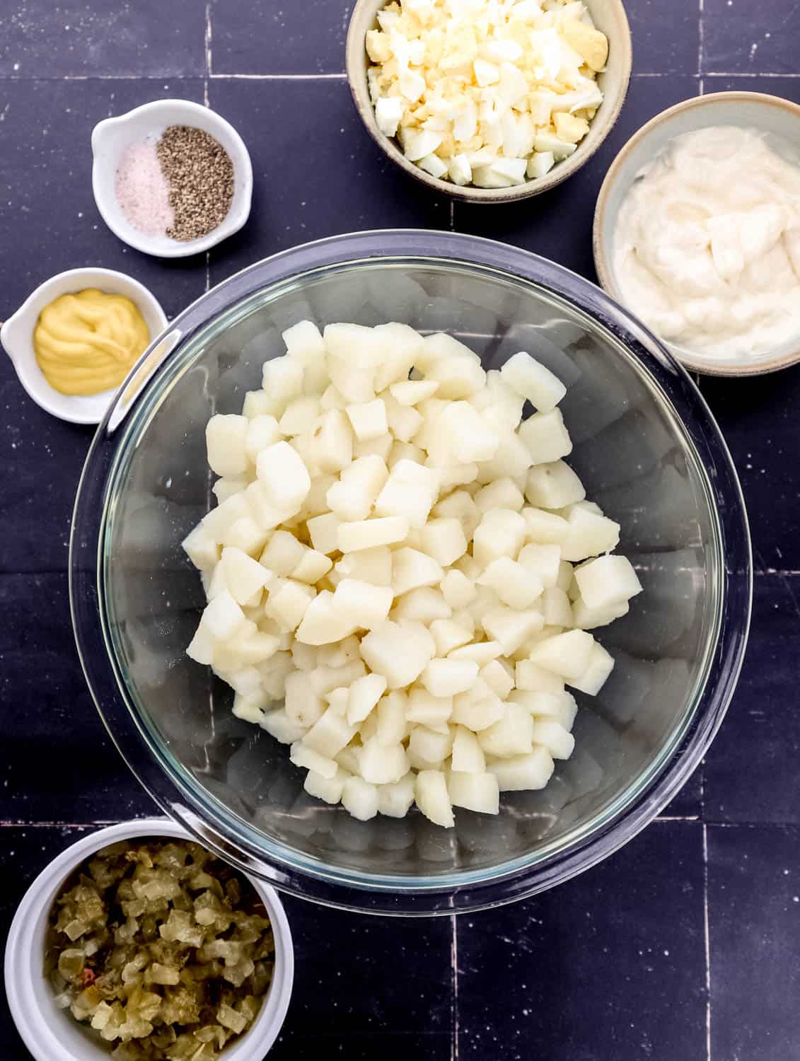 Overhead view of ingredients needed to make potato salad in separate bowls on black tile surface. 