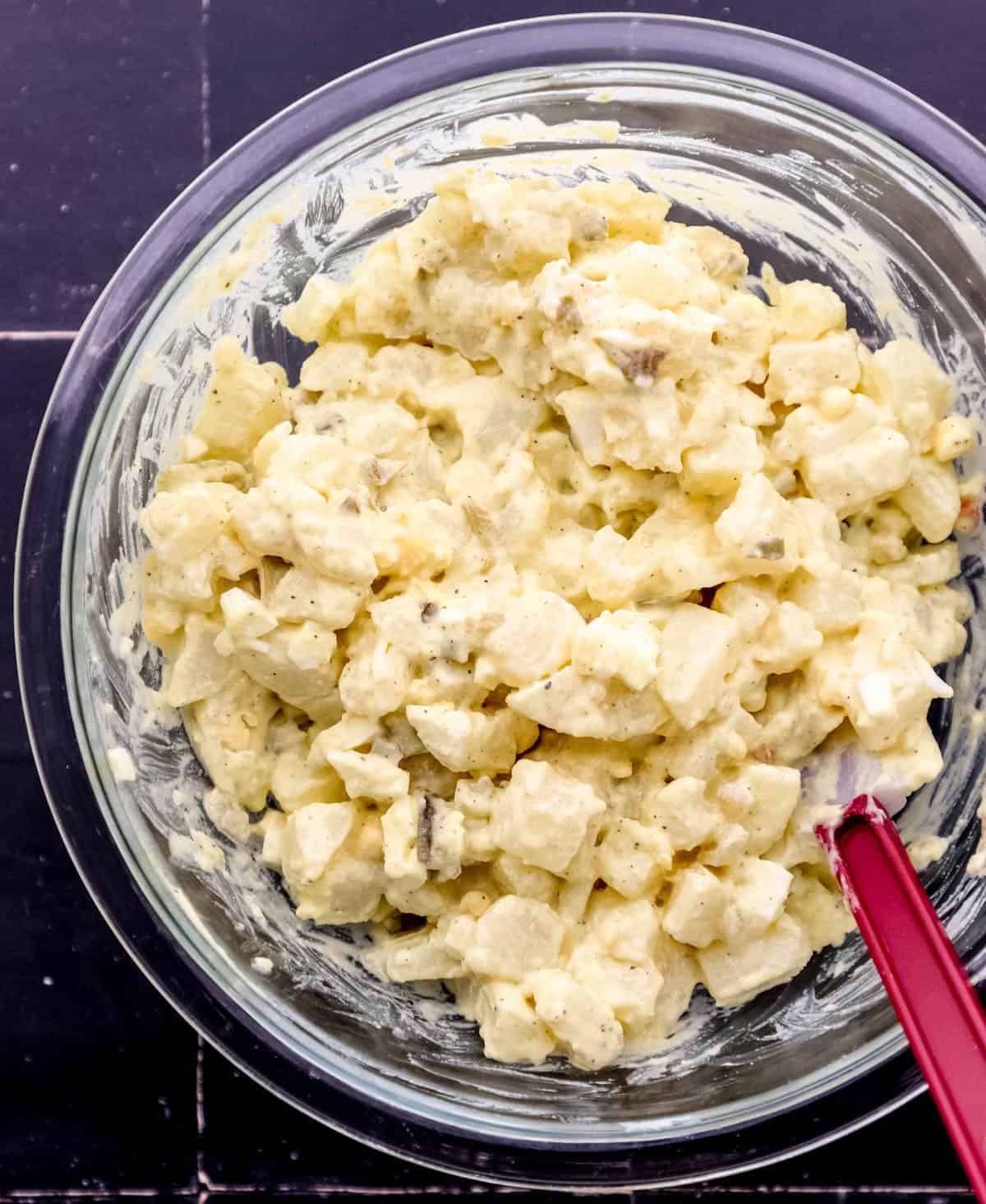 Overhead view of combined salad ingredients in glass mixing bowl with spatula. 