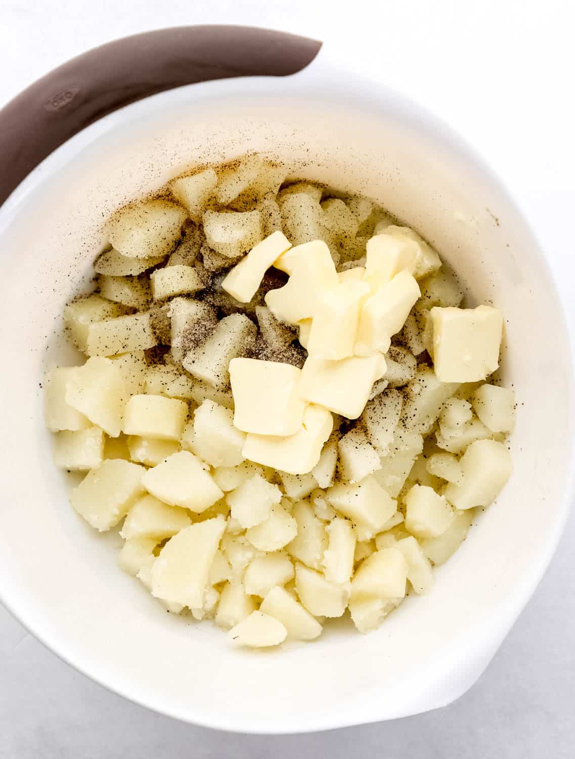 Potatoes, butter, salt, and ground black pepper in large white mixing bowl. 