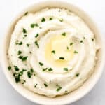 Overhead view of mashed potatoes in white bowl topped with butter and parsley on grey surface.