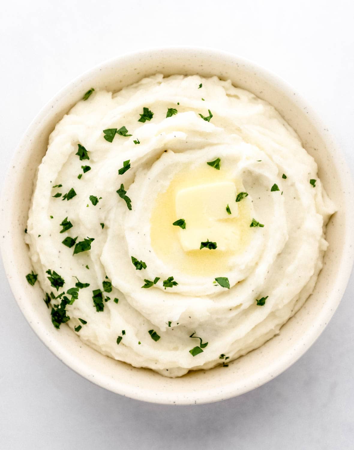 Overhead view of mashed potatoes topped with butter and parsley in a white bowl on grey surface.