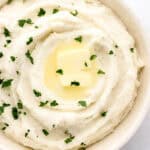Close up overhead view of potatoes in white bowl topped with butter and chopped parsley.