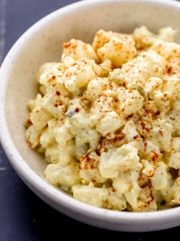 Close up side view of potato salad in white bowl on black tile surface.