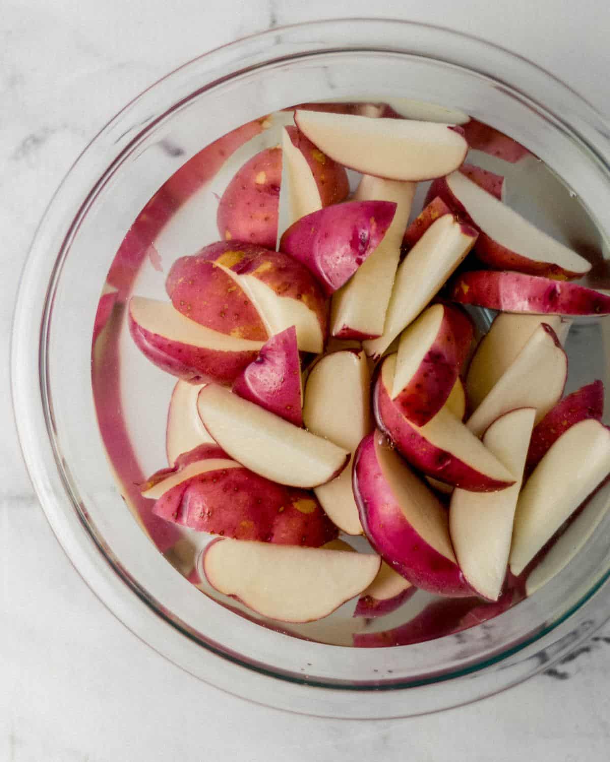 Sliced red potatoes in glass mixing bowl with cold water on marble surface. 