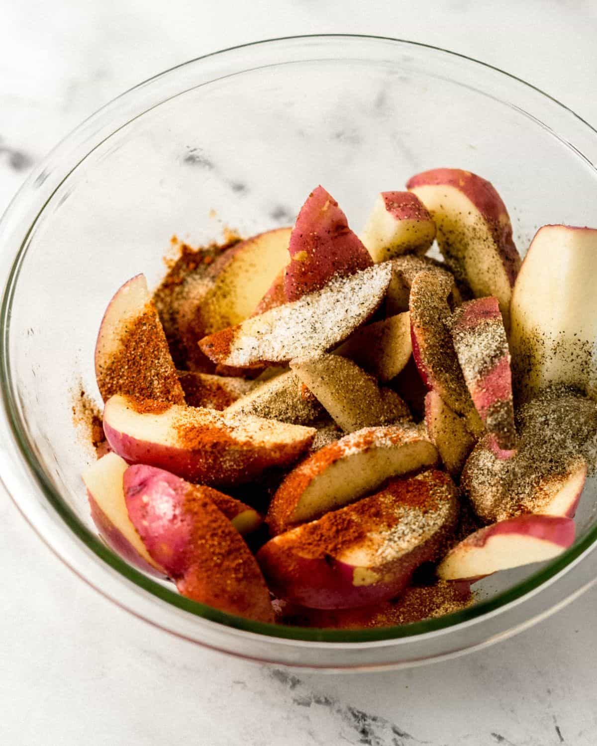 Potatoes topped with seasoning in glass bowl on marble surface. 