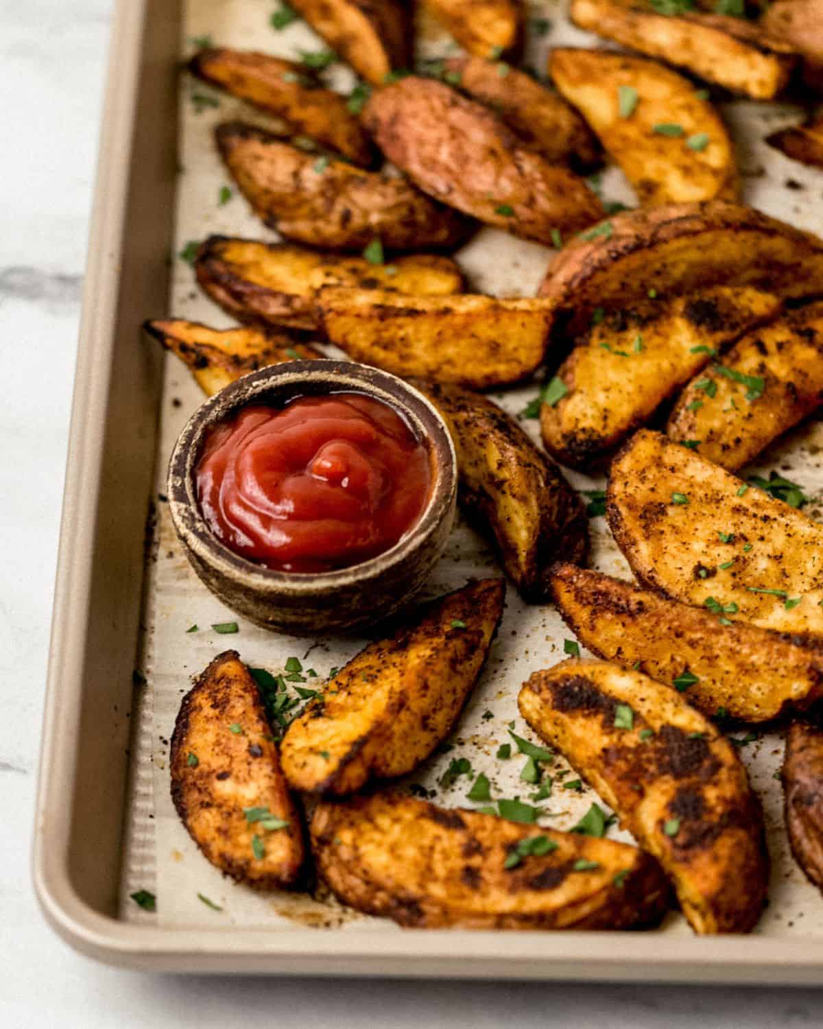 Close up front view of potato wedges on parchment lined baking sheet with small wooden bowl of ketchup. 