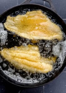 Two pieces of fish frying in oil in cast iron skillet.